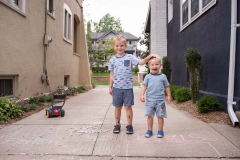 During a family documentary photo session two boys stand beside driveway chalk drawings and laugh