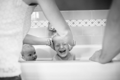 Family documentary photograph of a boy laughing as mother scrubs his head during a bath