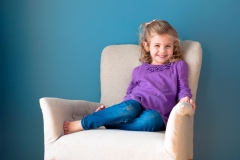 A cute young girl poses in a white chair against a stunning blue wall