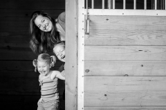 A mom and her two girls peer around a door and laugh brightly in this black and white family lifestyle portrait