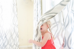 Wearing a bright orange dress a young girl plays peekaboo in window curtains for a lifestyle portrait