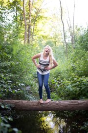 A senior girl laughs standing on a log over a stream in natural Grand Rapids senior portraits