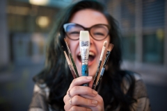 Artistic senior girl laughs as she holds up paint brushes for senior portraits in Grand Rapids