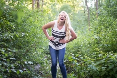 A senior girl laughs standing on a log over a stream in natural Grand Rapids senior portraits