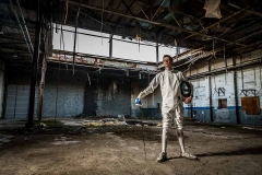 Grand Rapids senior guy in fencing outfit poses in a gritty urban warehouse for senior pictures