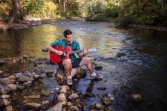 Senior guy plays guitar seated on a boulder in a West Michigan stream