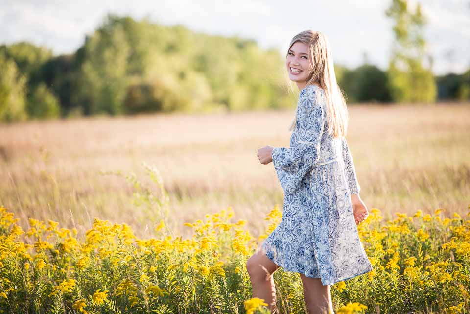 Grand Rapids girl in a dress smiles broadly as she walks through a fall West Michigan field for high school senior portraits
